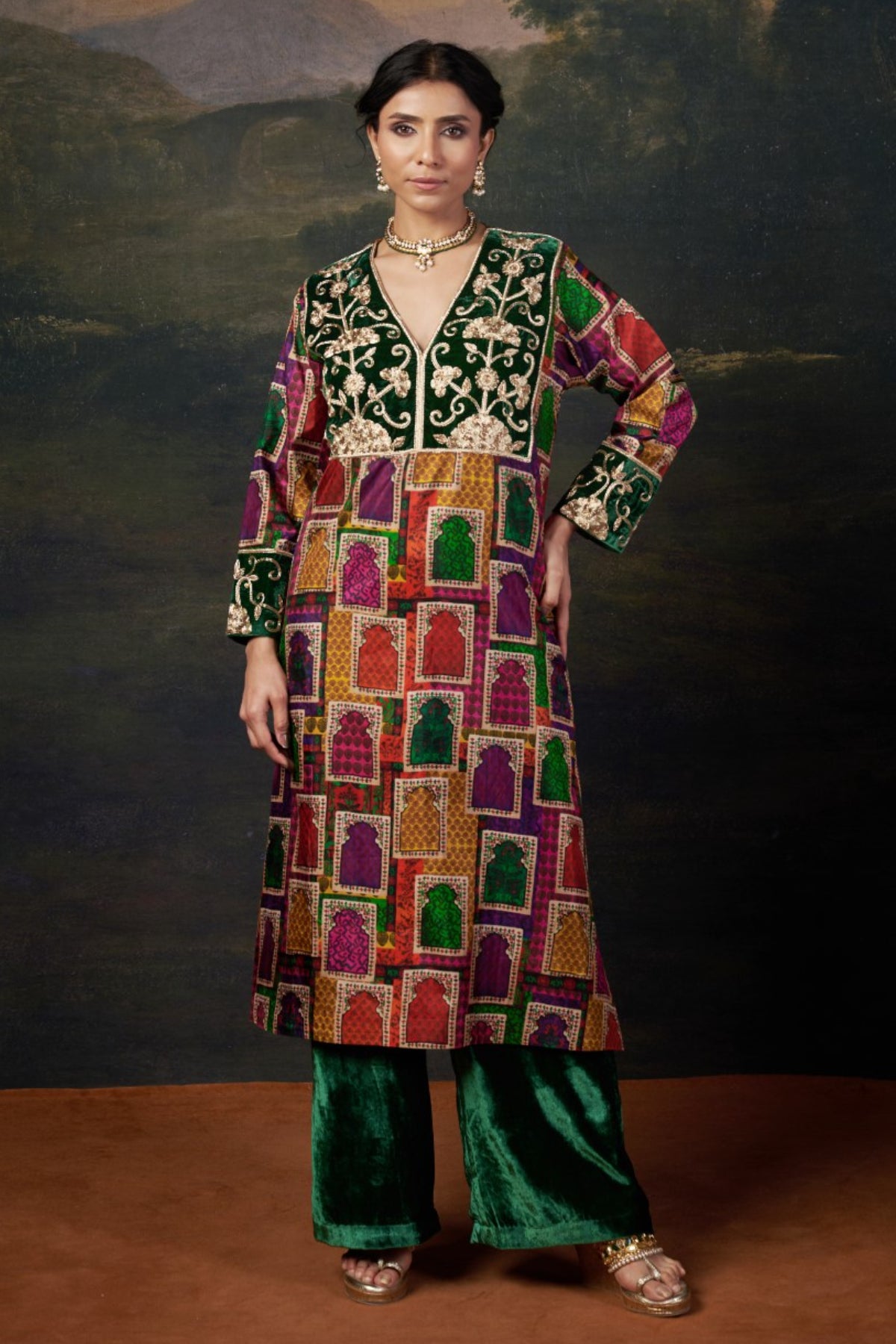 Woman wearing a colorful traditional outfit with a patterned top and green pants against a dark background.